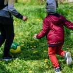 Two children enjoy a game of soccer in a sunny, grassy park area surrounded by dandelions.